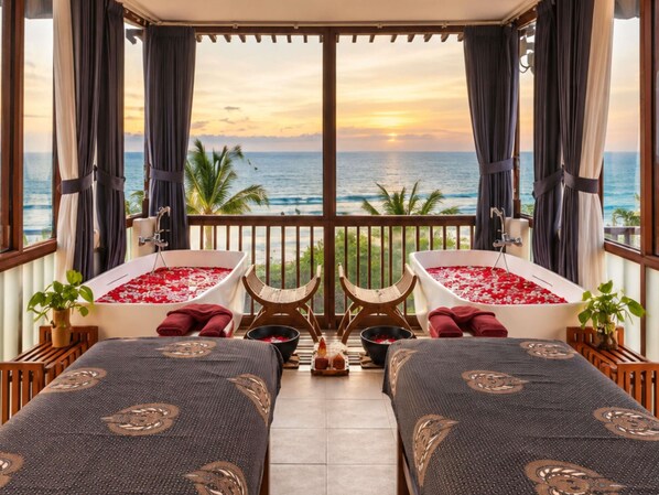 A room with two large tubs filled with rose petals, a bed with a patterned bedspread, and a view of the ocean at sunset.