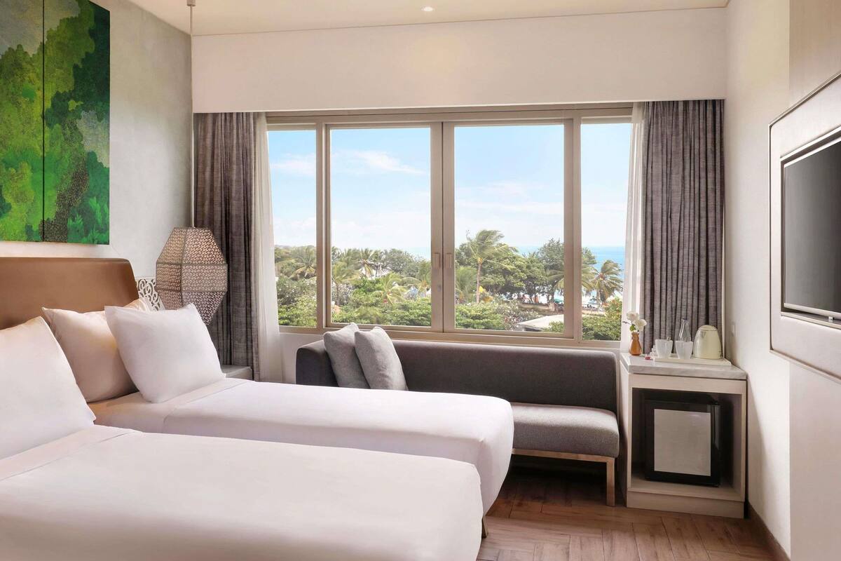 A hotel room with two beds, a sofa, a TV, and a large window with a view of greenery and palm trees.