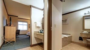 A hotel room with a wooden desk, a chair, a small kitchenette, a bathroom with a sink and mirror, and a window. - Dimond Center Hotel (Anchorage)