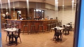 A restaurant with a bar area, multiple tables and chairs, and a counter with a person behind it. - Dimond Center Hotel (Anchorage)