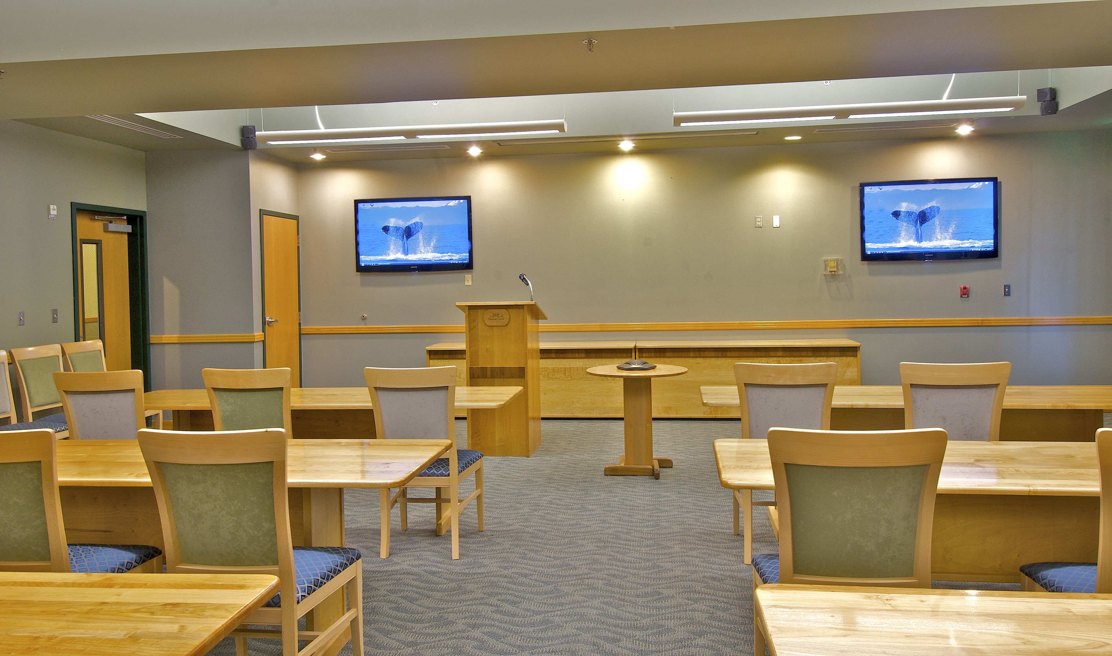 A conference room with multiple tables and chairs, two large screens on the wall, and a podium.