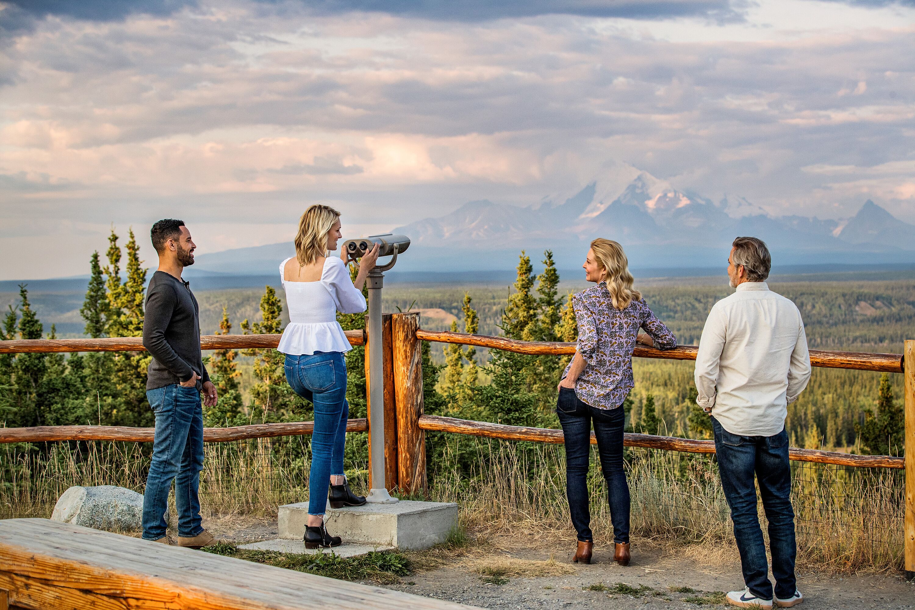 Een groep mensen staat op een houten terras met uitzicht op een bergachtig landschap.