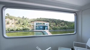 Une pièce dotée d’une grande fenêtre donnant sur une étendue d’eau et sur un bâtiment à l’architecture moderne. On y trouve des chaises blanches et une petite table.