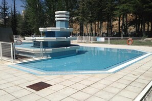 A rectangular swimming pool with a central fountain structure, surrounded by a fence and trees in the background. - Hotel Nayade (Vegas de Matute)