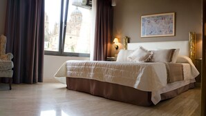 Una habitación de hotel con una cama, una silla, un ventanal con cortinas, una lámpara y un cuadro en la pared.