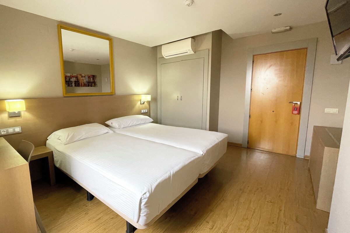 A hotel room with a double bed, a desk, a chair, a wardrobe, a mirror, and a door.
