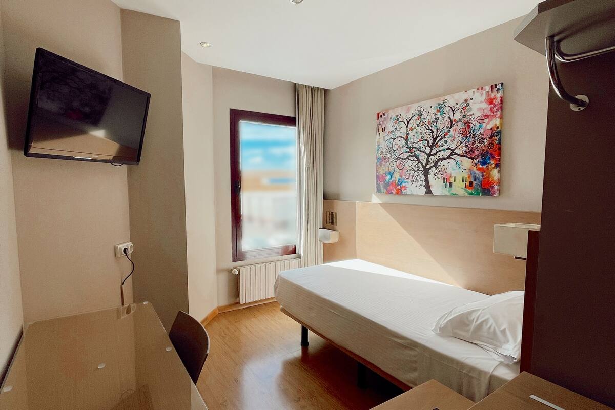 A hotel room with a bed, a desk, a television, a window with curtains, and a painting on the wall.