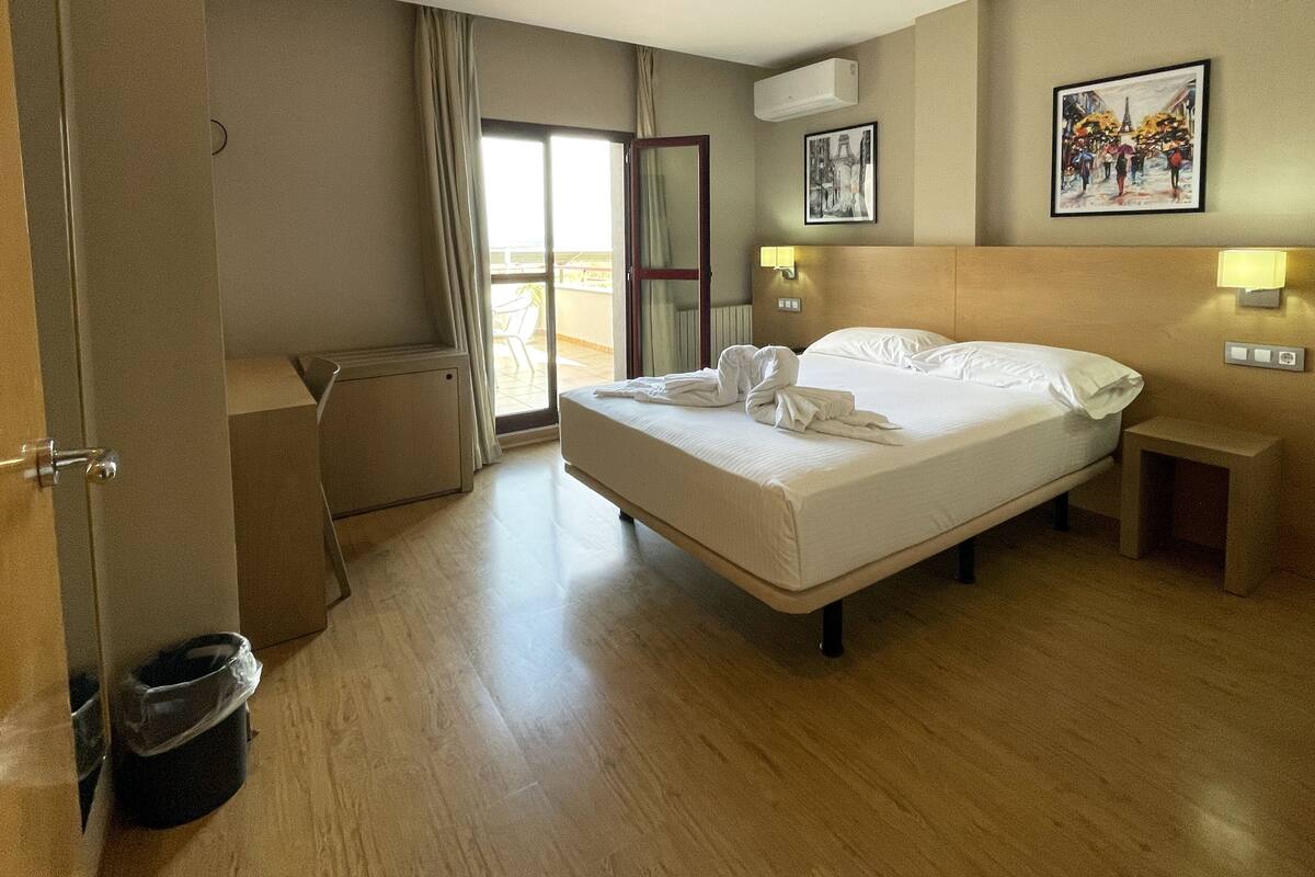 A hotel room with a bed, a desk, a chair, a trash bin, and a window with curtains.