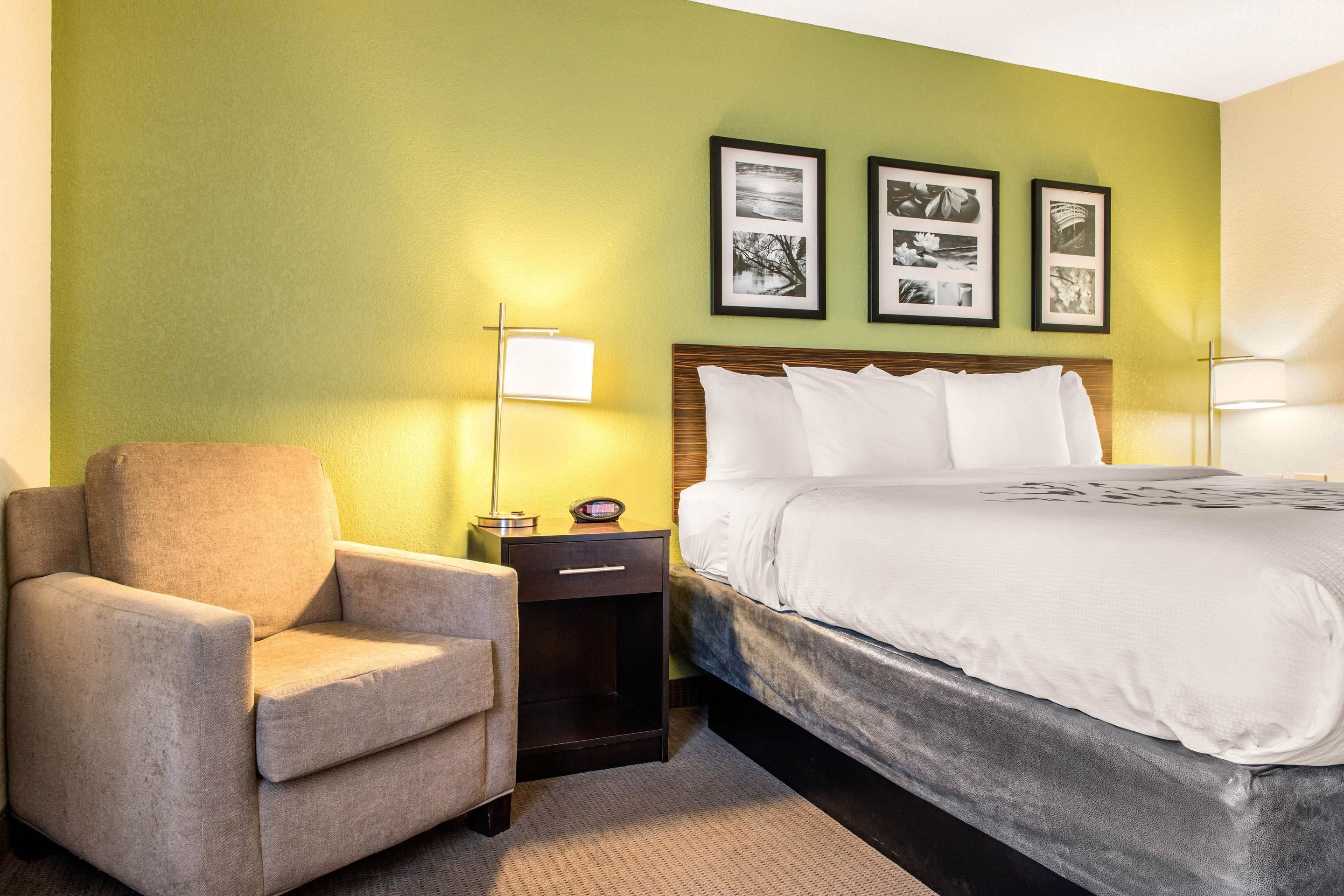 A hotel room with a bed, a chair, a nightstand, and a wall with framed pictures.