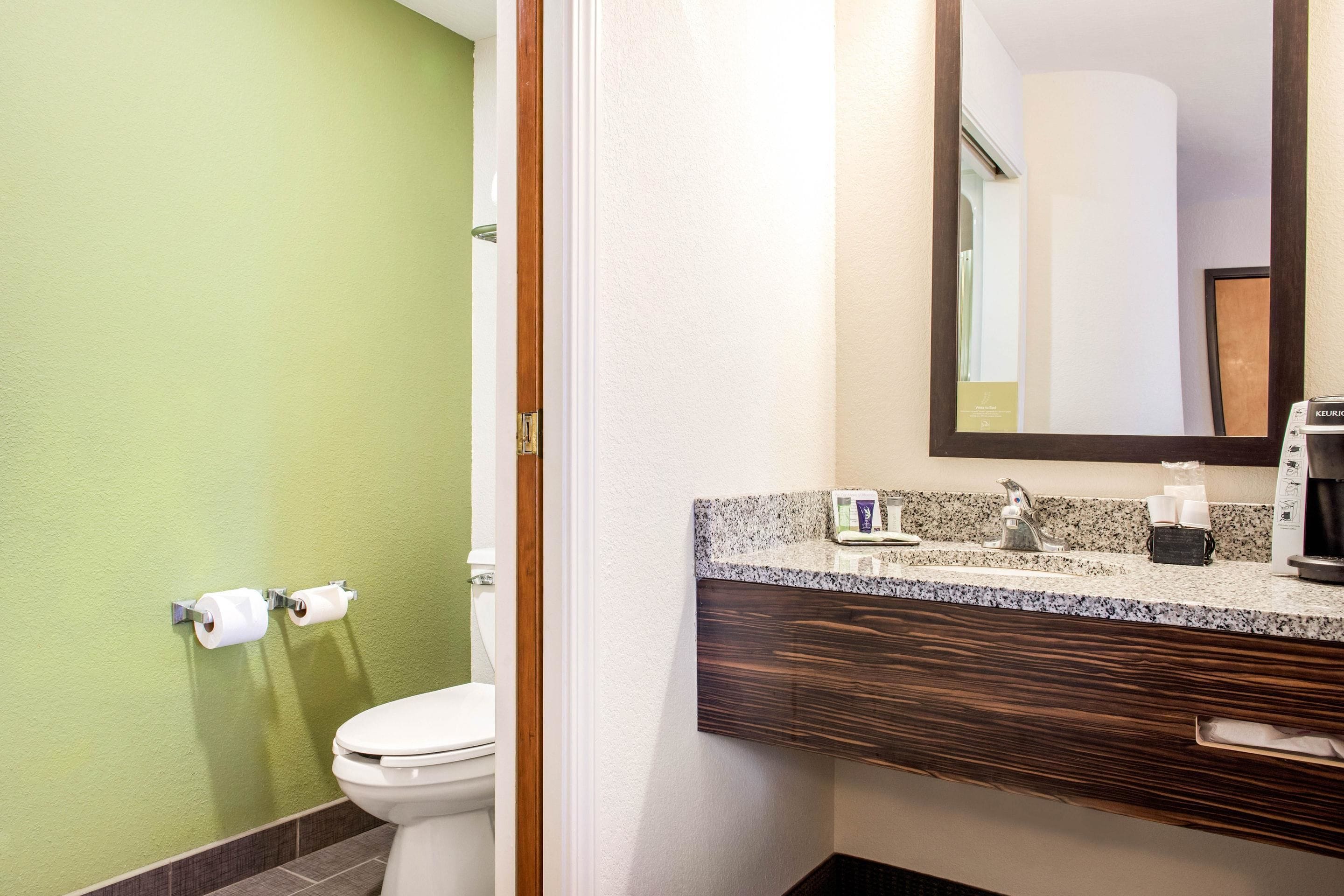 A bathroom with a green wall, a white toilet, a wooden vanity with a mirror, and a sink with a coffee maker.