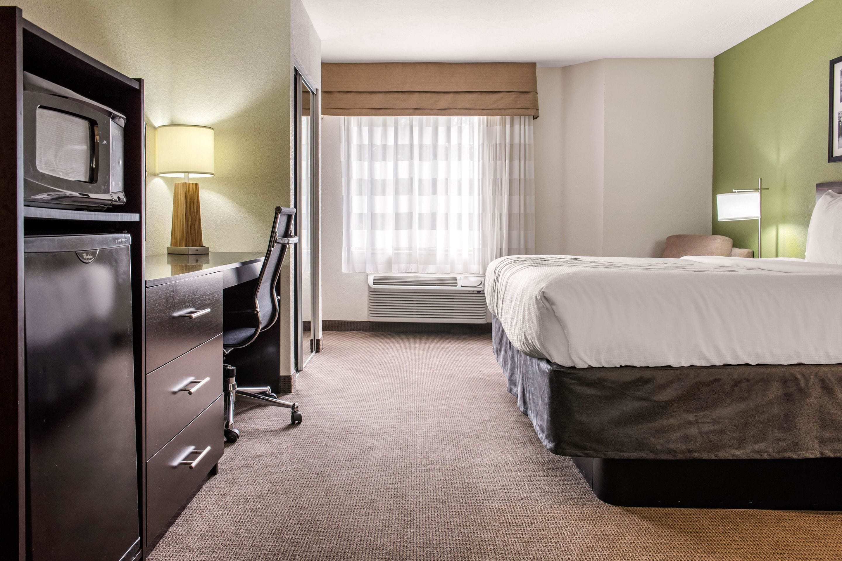 Ein Hotelzimmer mit Bett, Schreibtisch mit Lampe, Stuhl, Kommode und einem Fenster mit Vorhängen.