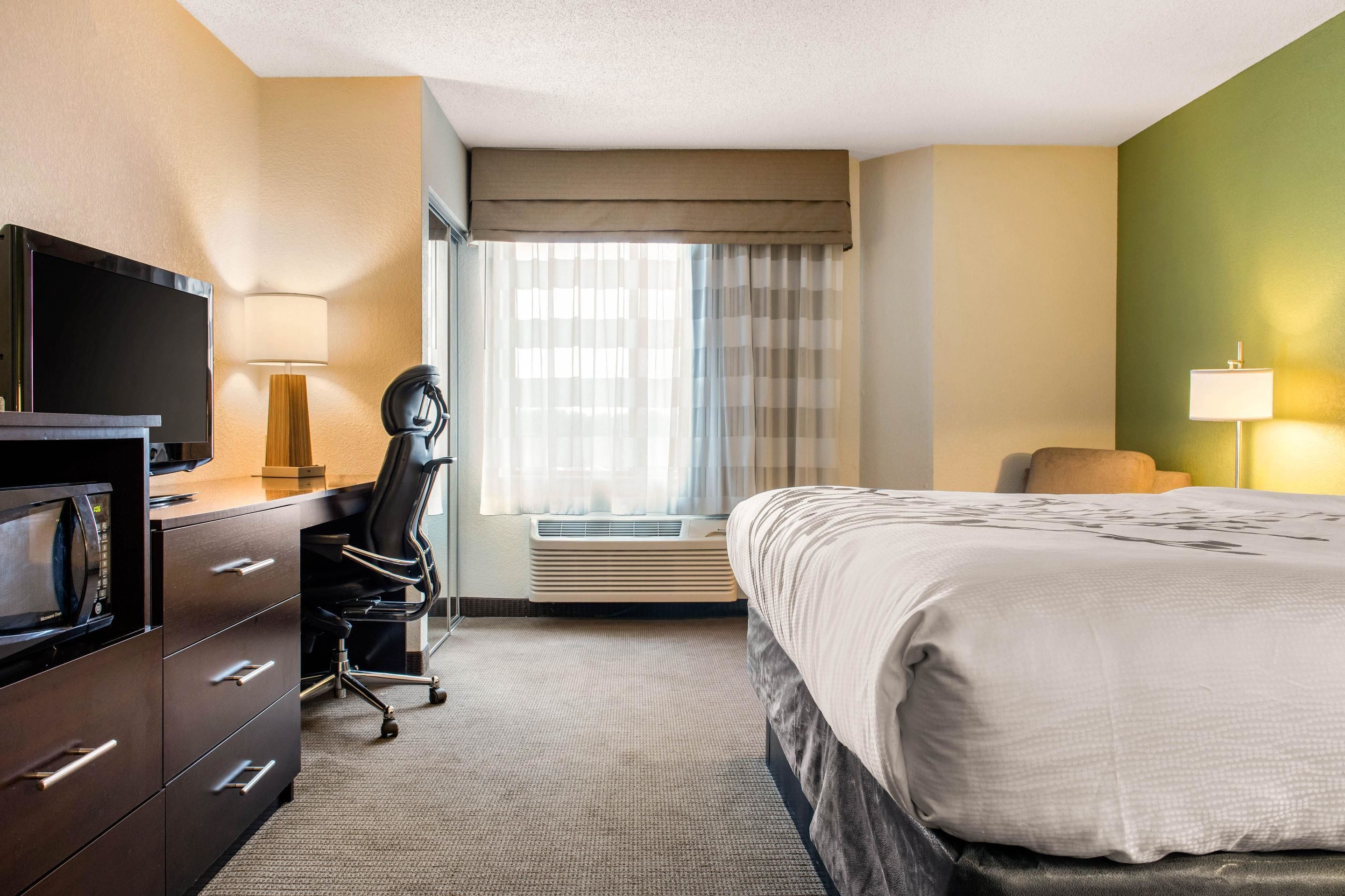 A hotel room with a bed, a desk with a computer, a chair, a TV, and a window with curtains.
