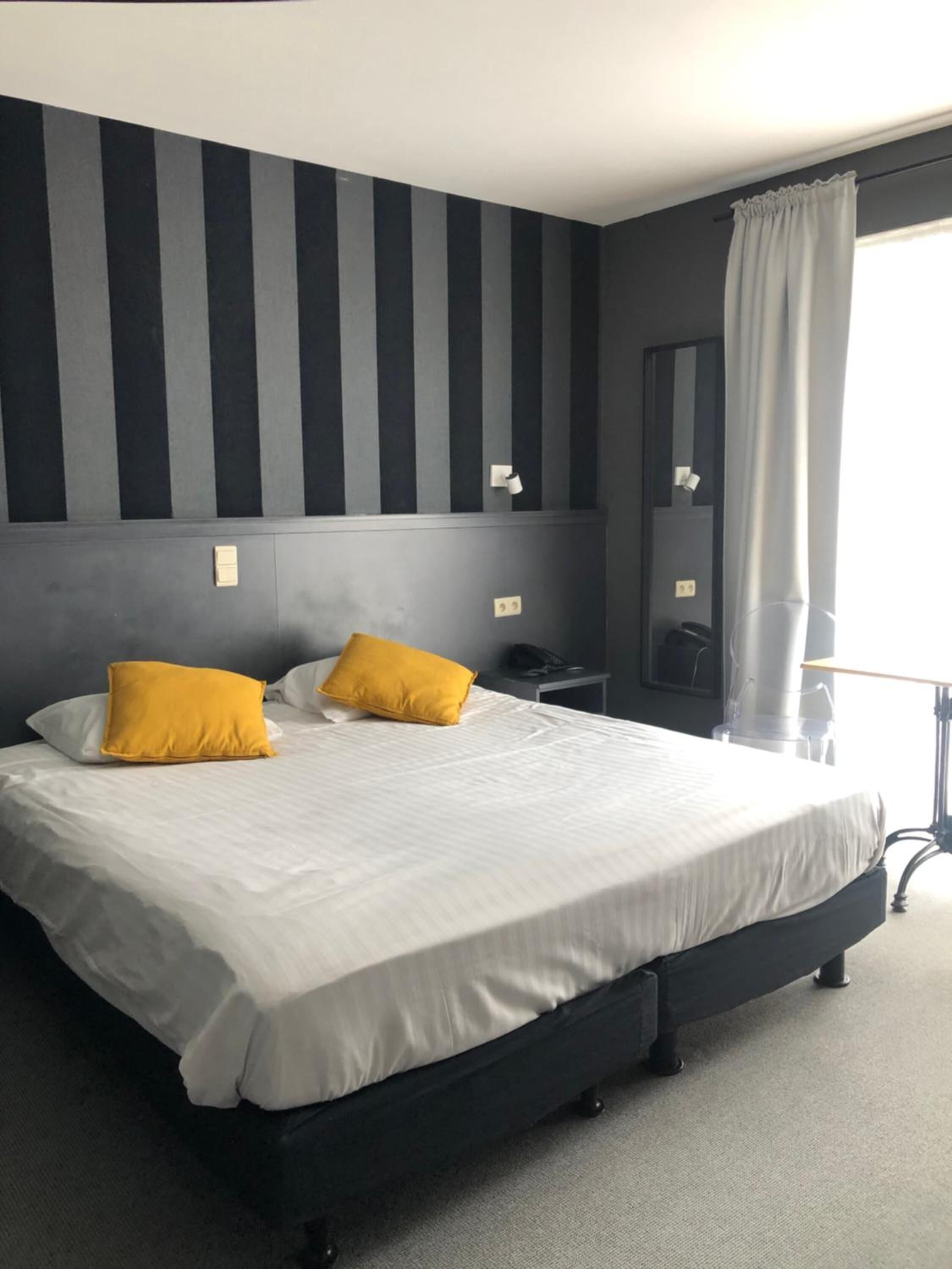 Habitación de hotel con una cama grande, dos almohadas, una mesita de noche, una lámpara y un ventanal con cortinas.