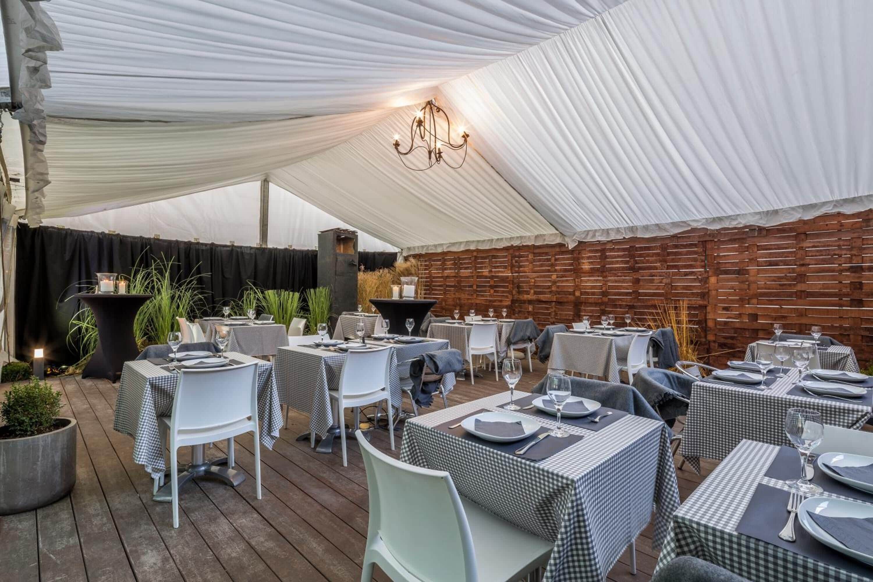 Een terras met tafels gedekt voor gasten, een witte tent erboven en een bakstenen muur op de achtergrond.