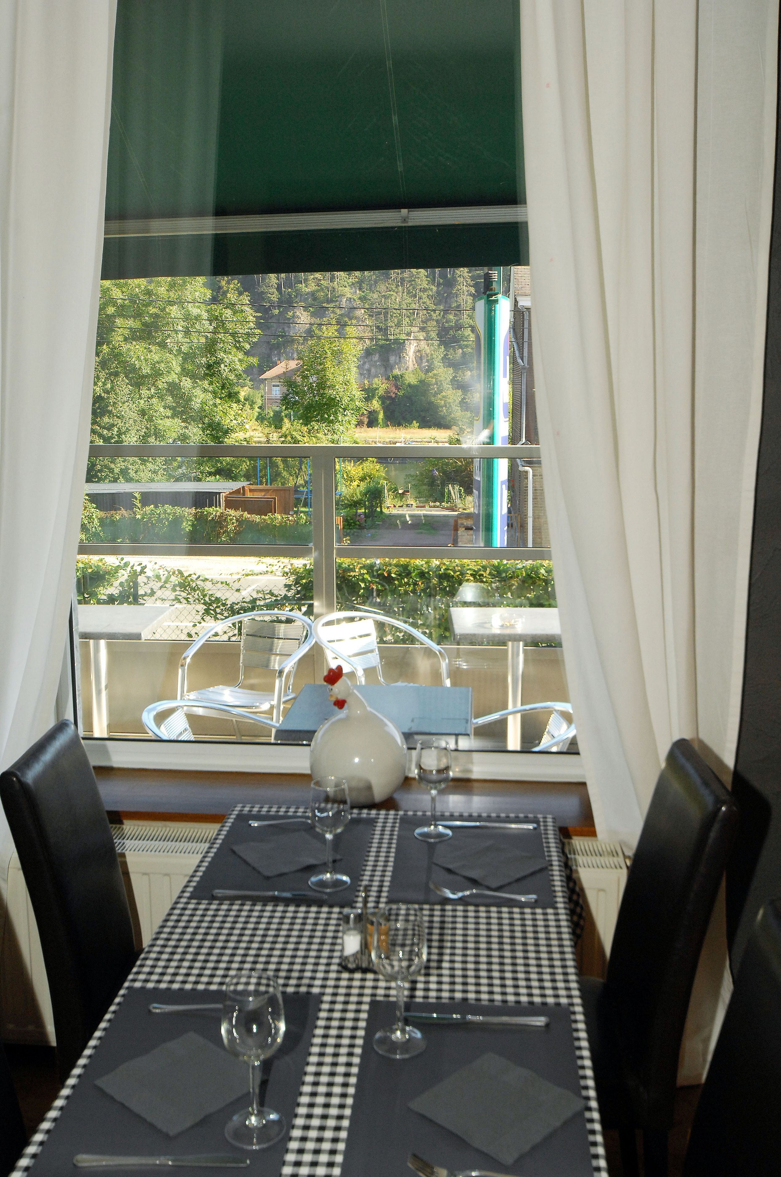 Une table à manger dressée pour deux personnes, avec une nappe à carreaux, deux verres à vin et des couverts. La table est placée près d’une fenêtre donnant sur un balcon et sur la verdure à l’extérieur.
