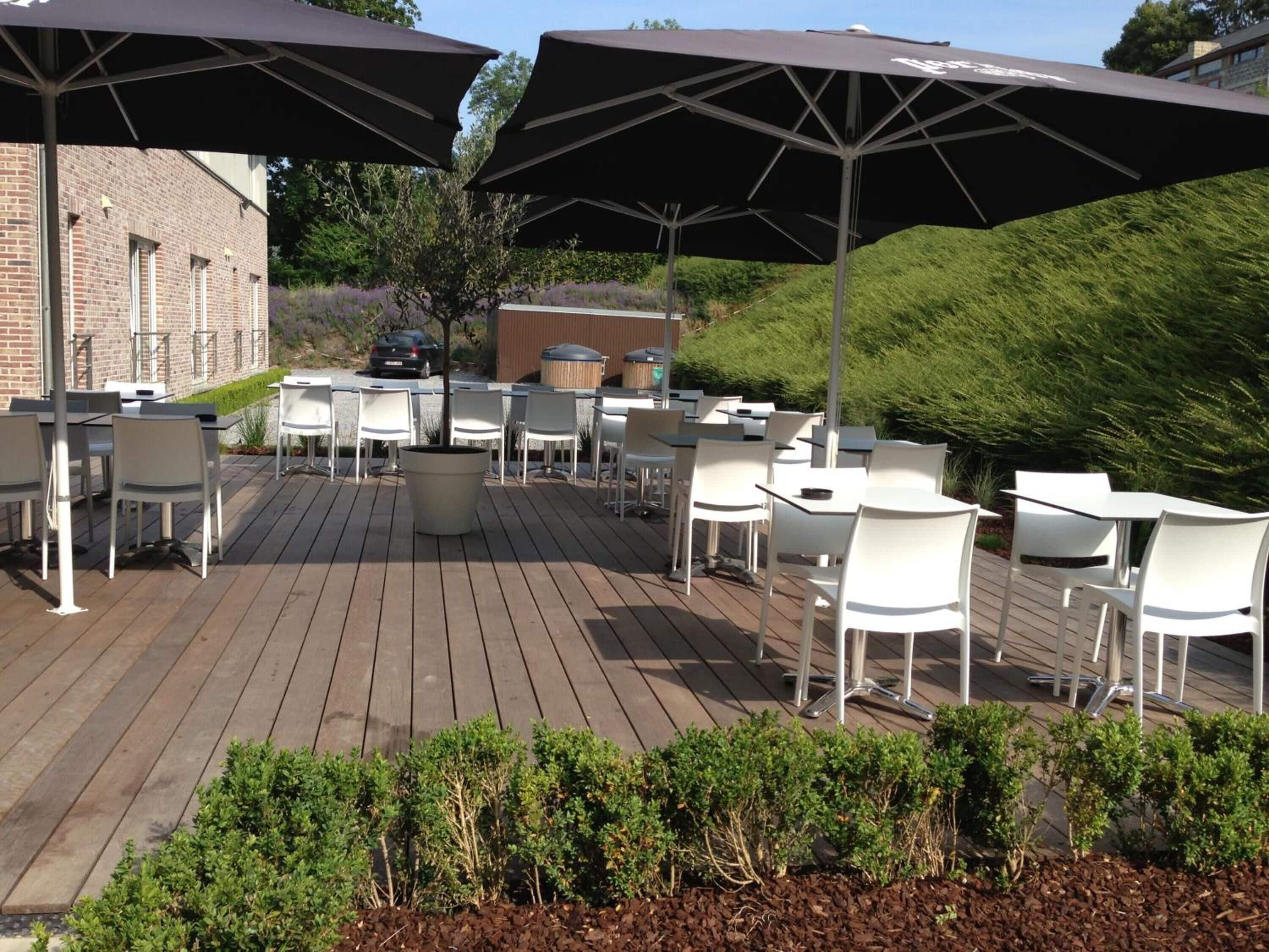 Een terras buiten met witte tafels en stoelen, grote parasols en een bakstenen gebouw op de achtergrond.