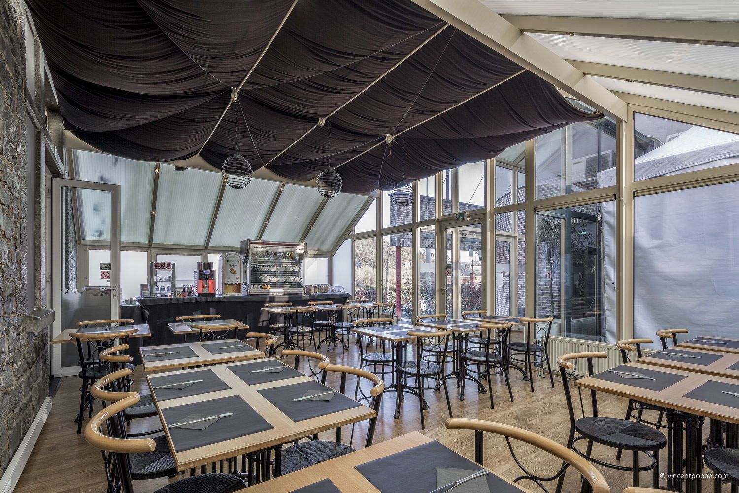 A modern cafe with a high ceiling, large windows, and a central counter with a coffee machine. Tables are arranged in a U-shape around the counter.