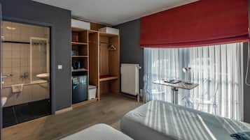 A modern hotel room with a wooden floor, a bed with striped bedding, a desk with a chair, a wardrobe, and a bathroom with a shower and toilet.