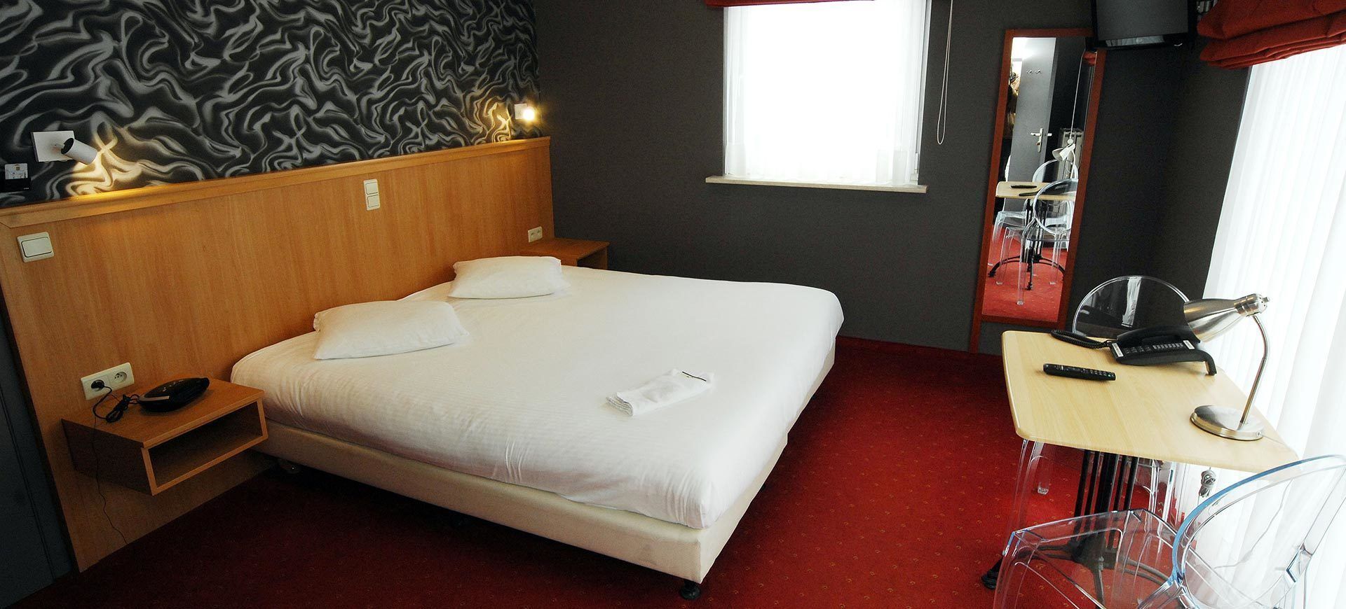 A hotel room with a bed, bedside table, and a desk with a chair.