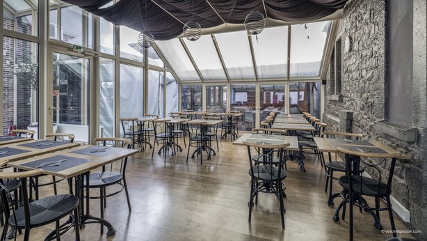A spacious indoor dining area with high ceilings, large windows, and a stone wall. Tables and chairs are arranged for dining.