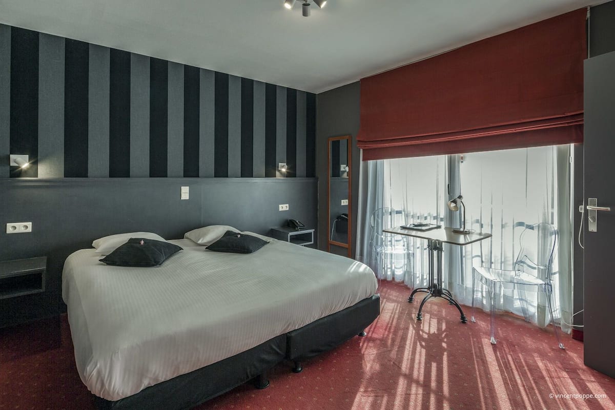 Een hotelkamer met een groot bed, een nachtkastje, een bureau met een stoel en uitzicht op de stad door grote ramen.