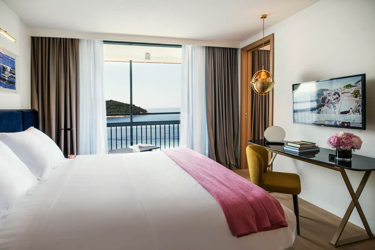 Quarto de hotel com uma cama grande, uma escrivaninha com cadeira, televisão e vista para o mar.