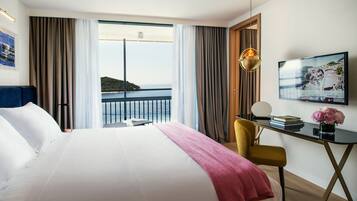 Quarto de hotel com uma cama grande, uma escrivaninha com cadeira, televisão e vista para o mar.