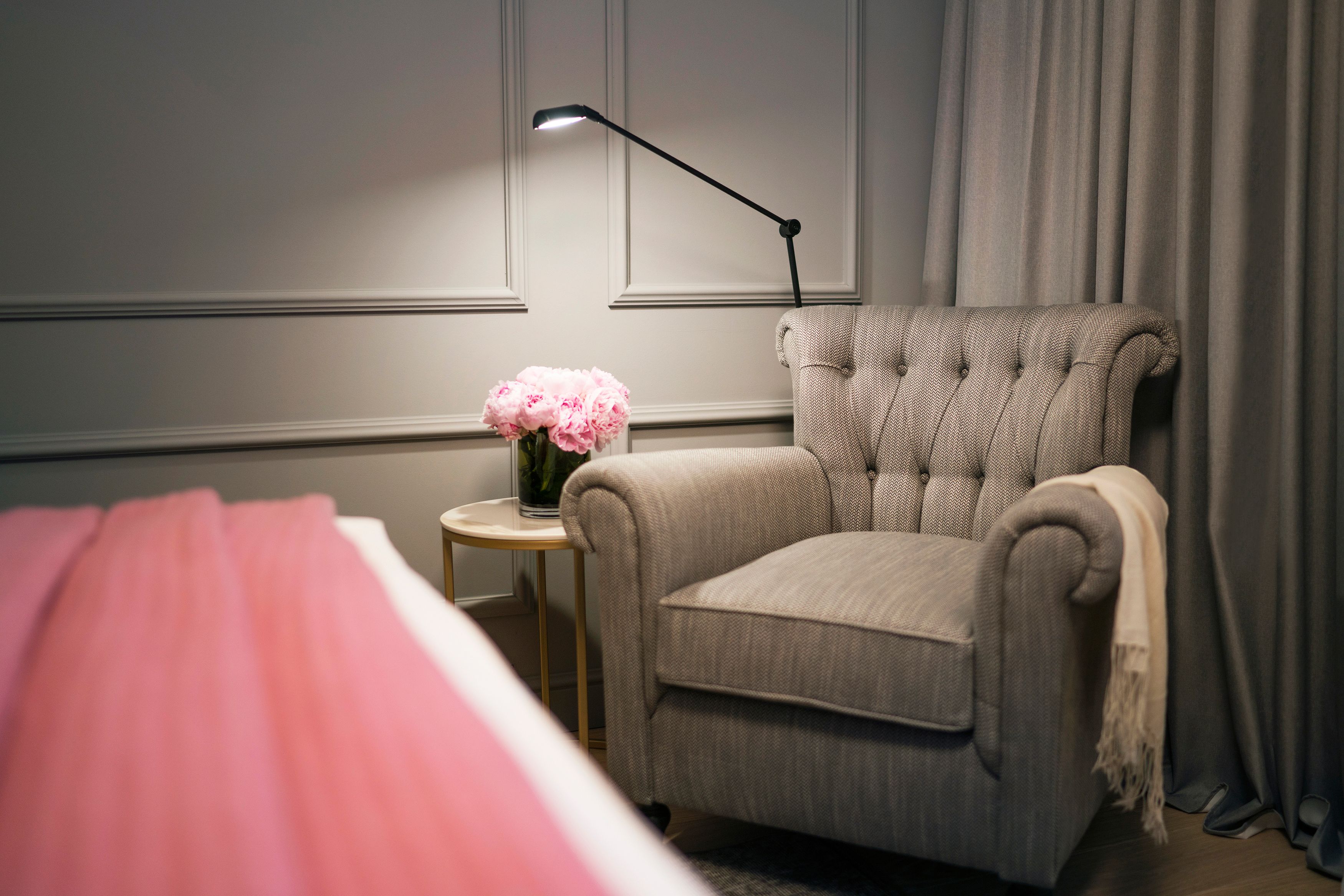 A room with a tufted armchair, a small side table with a vase of flowers, and a lamp.