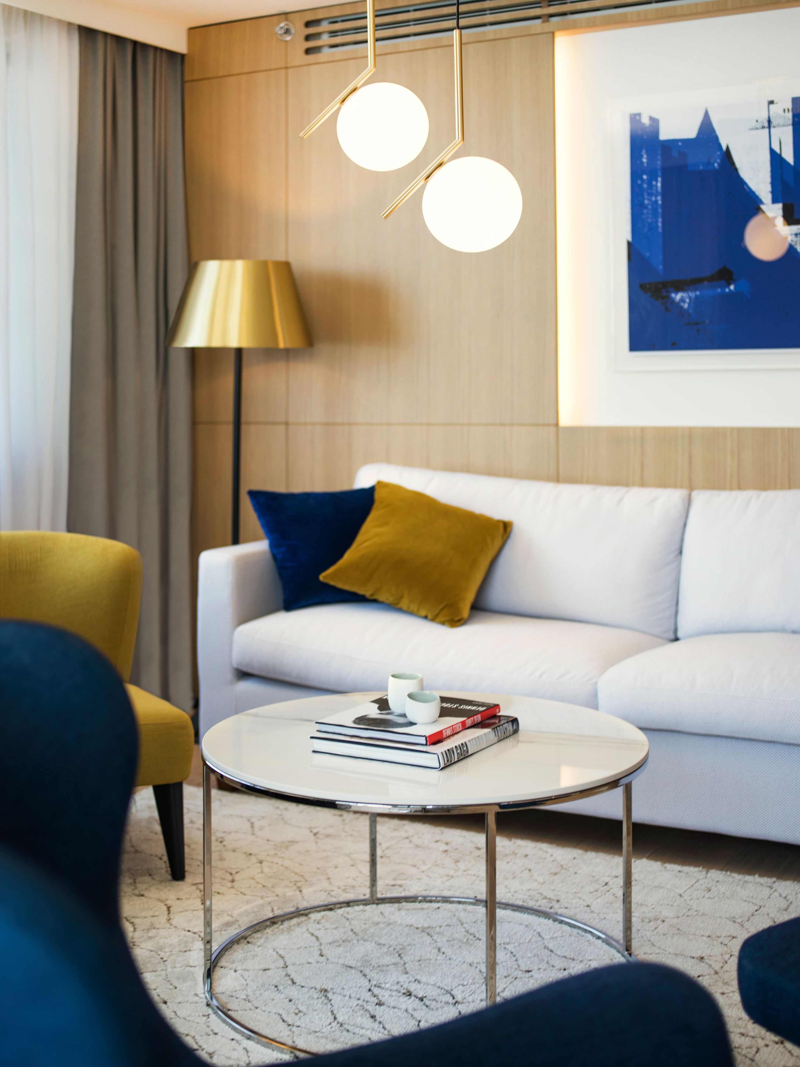 A modern living room with a white sofa, a round coffee table, and a yellow armchair. There is a lamp and a framed picture on the wall.