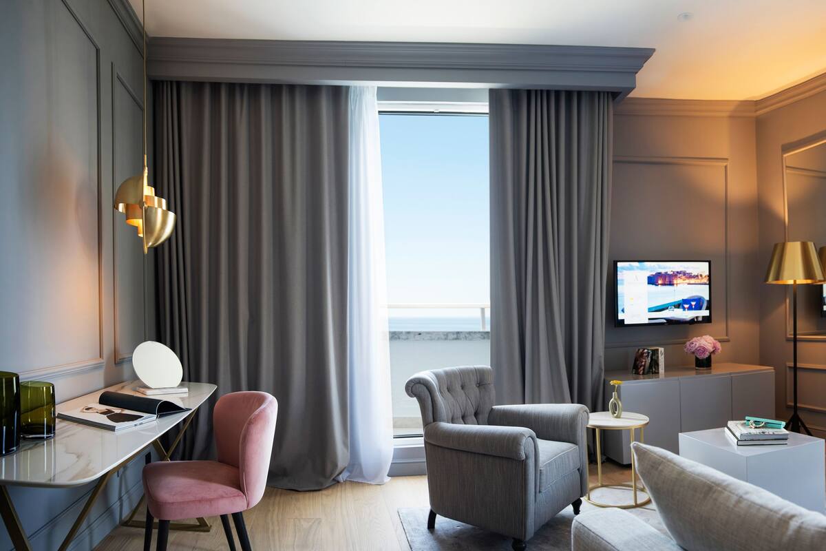 Quarto de hotel moderno com uma janela grande, uma escrivaninha com abajur, uma poltrona confortável, um sofá e uma TV de tela plana.