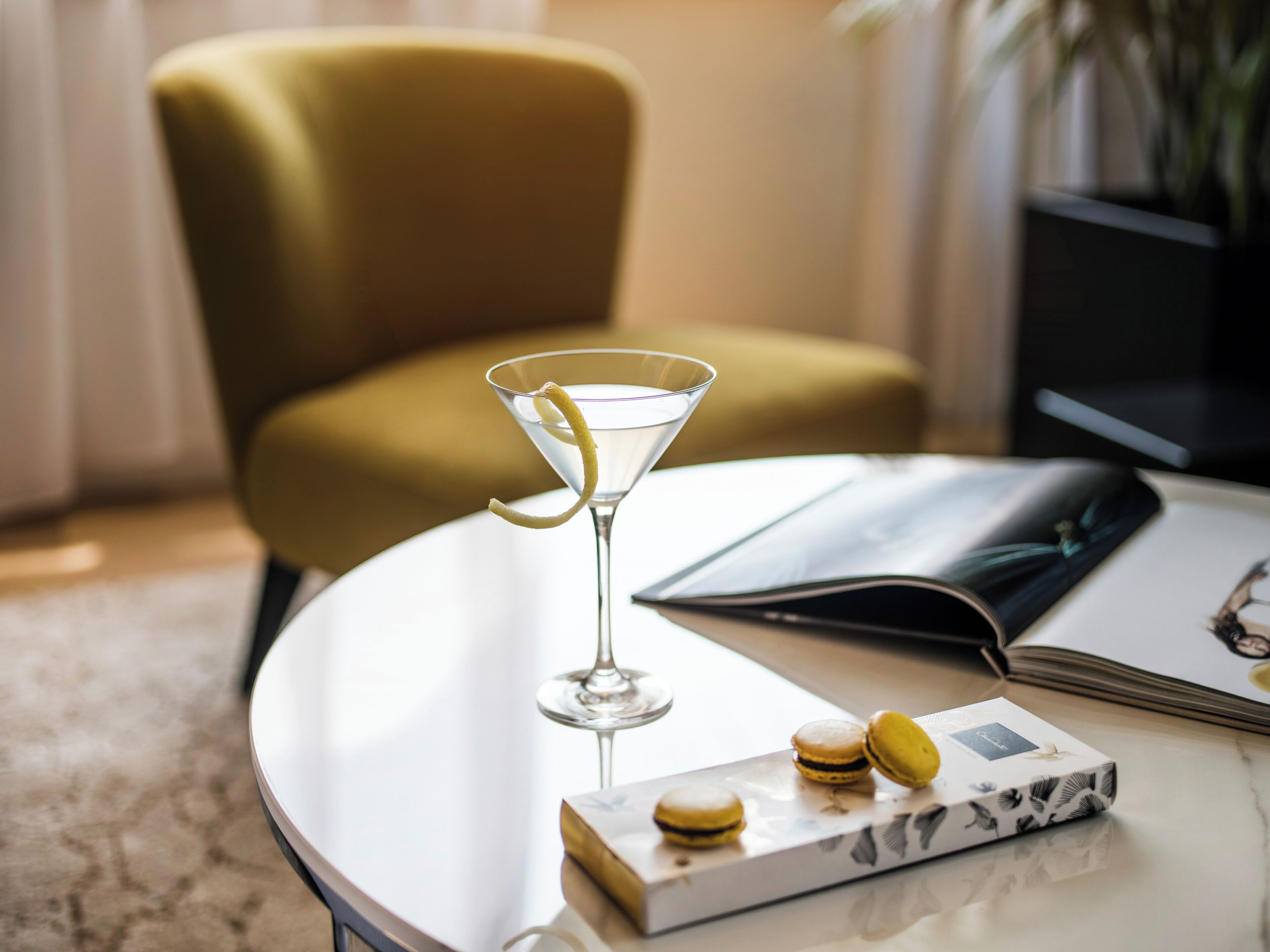 Un verre à martini orné d’une tranche de citron, posé sur une table ronde, à côté d’un livre et d’une boîte de macarons.