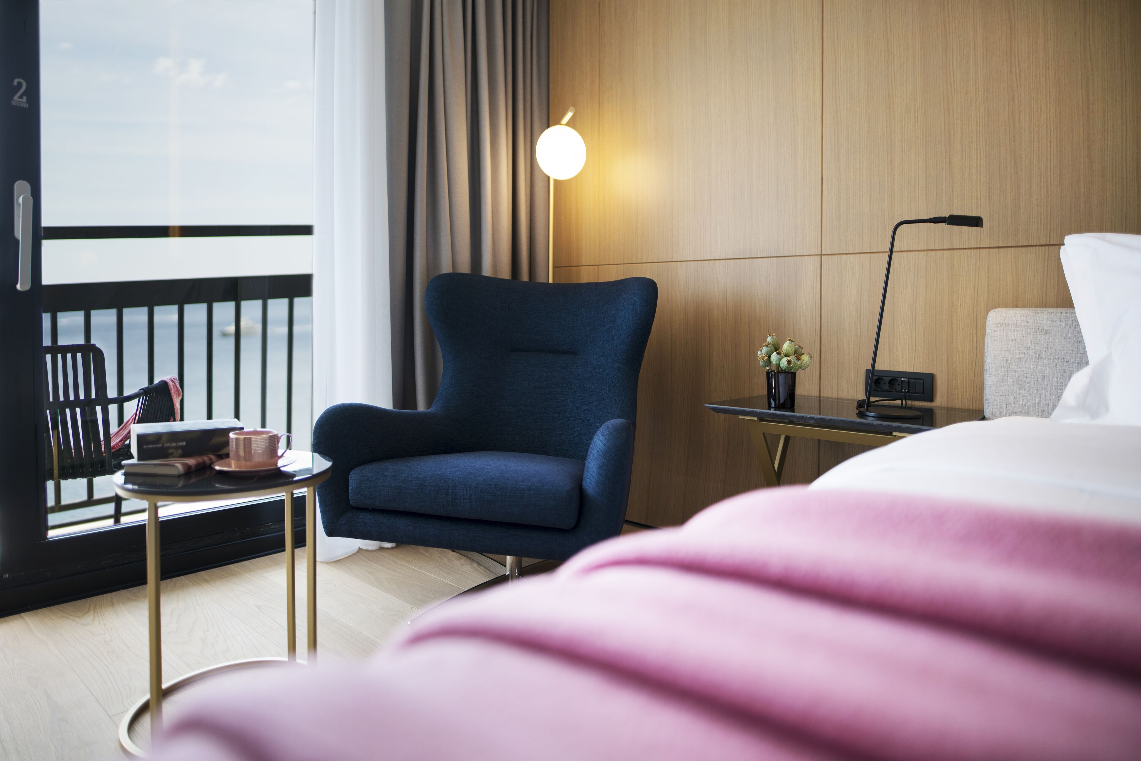 A hotel room with a bed, a blue armchair, a small table with a cup and books, a lamp, and a view of the sea through a window.
