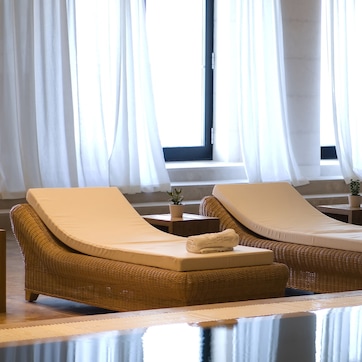 A room with two wicker lounge chairs with cushions, a small table with a plant, and a view of a building through large windows with white curtains.