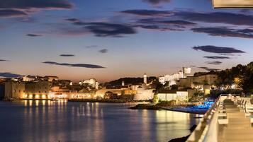 A coastal cityscape at dusk with illuminated buildings and a calm sea.