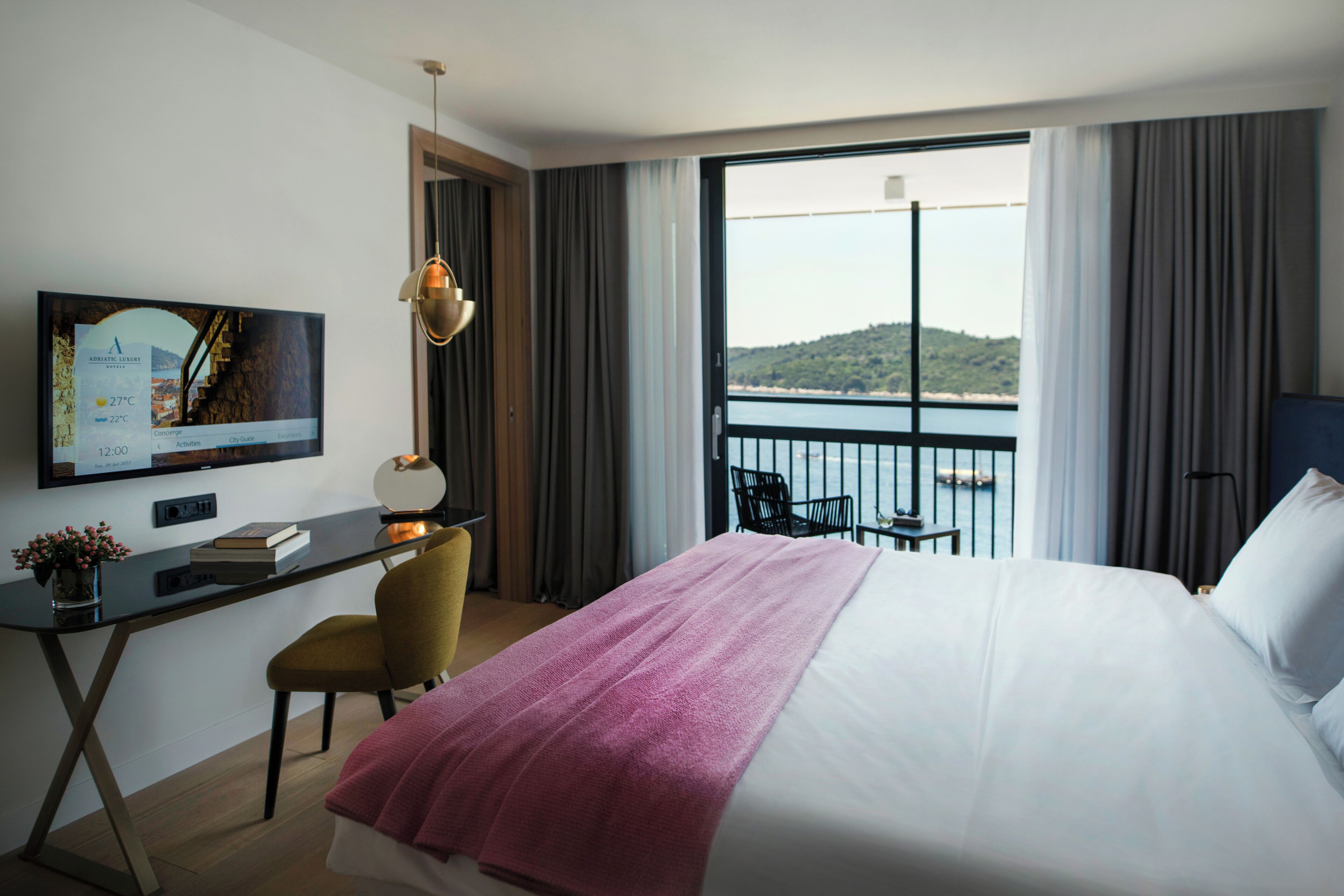 Habitación de hotel con cama, escritorio con computadora, silla, televisor en la pared y balcón con vista al mar.