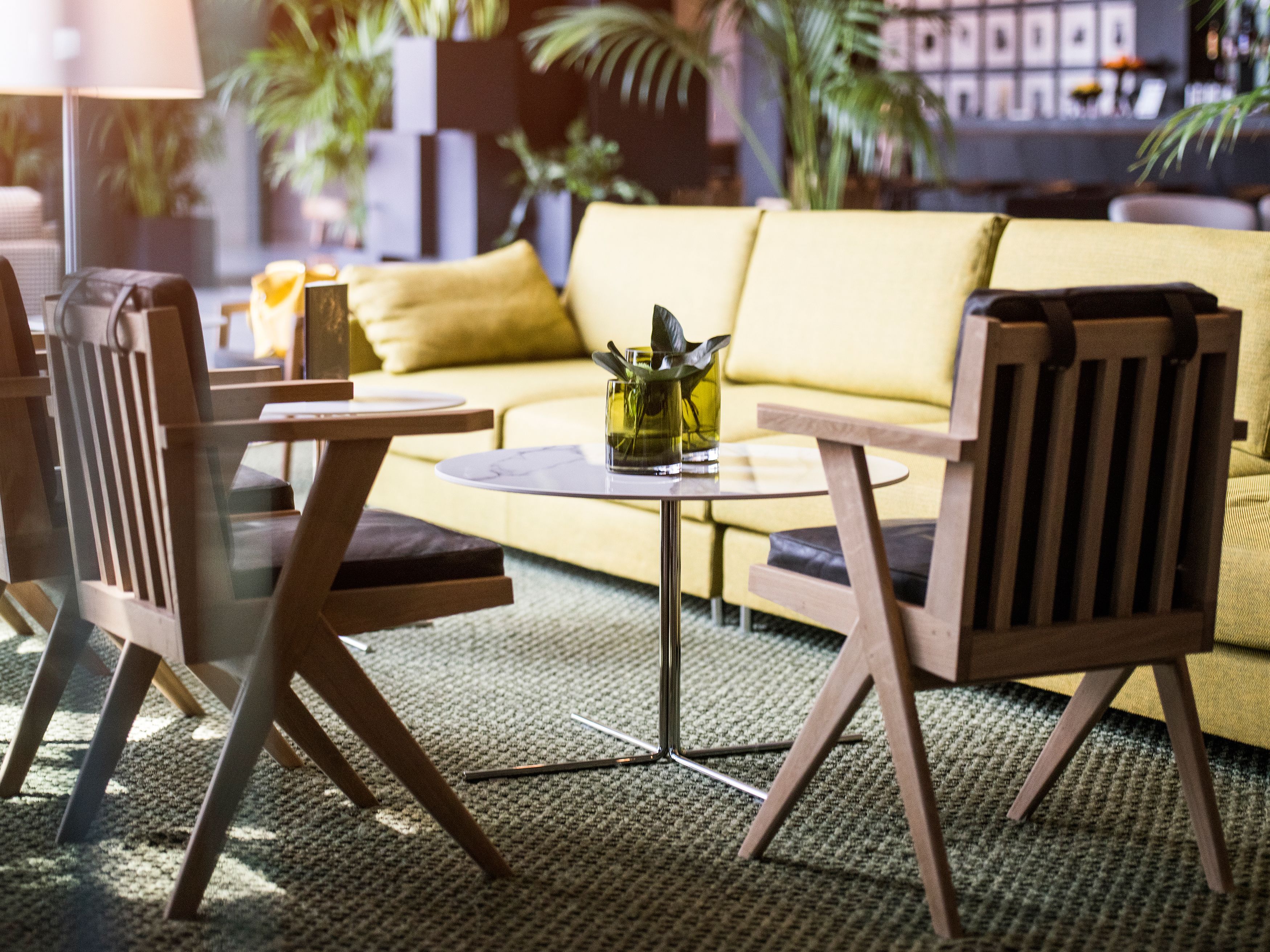 Uma sala de estar moderna com um sofá amarelo, cadeiras de madeira, uma mesa redonda pequena e um vaso com plantas verdes.