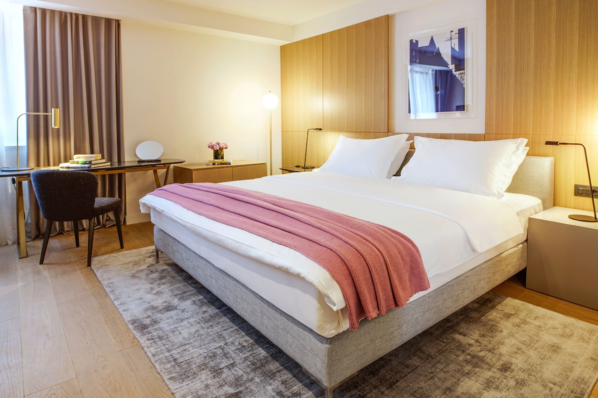 Quarto de hotel com uma cama grande, uma escrivaninha com cadeira, uma mesinha de apoio com abajur e um quadro na parede.