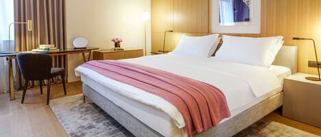 Una habitación de hotel con una cama grande, un escritorio con silla, una mesita con lámpara y un cuadro en la pared.