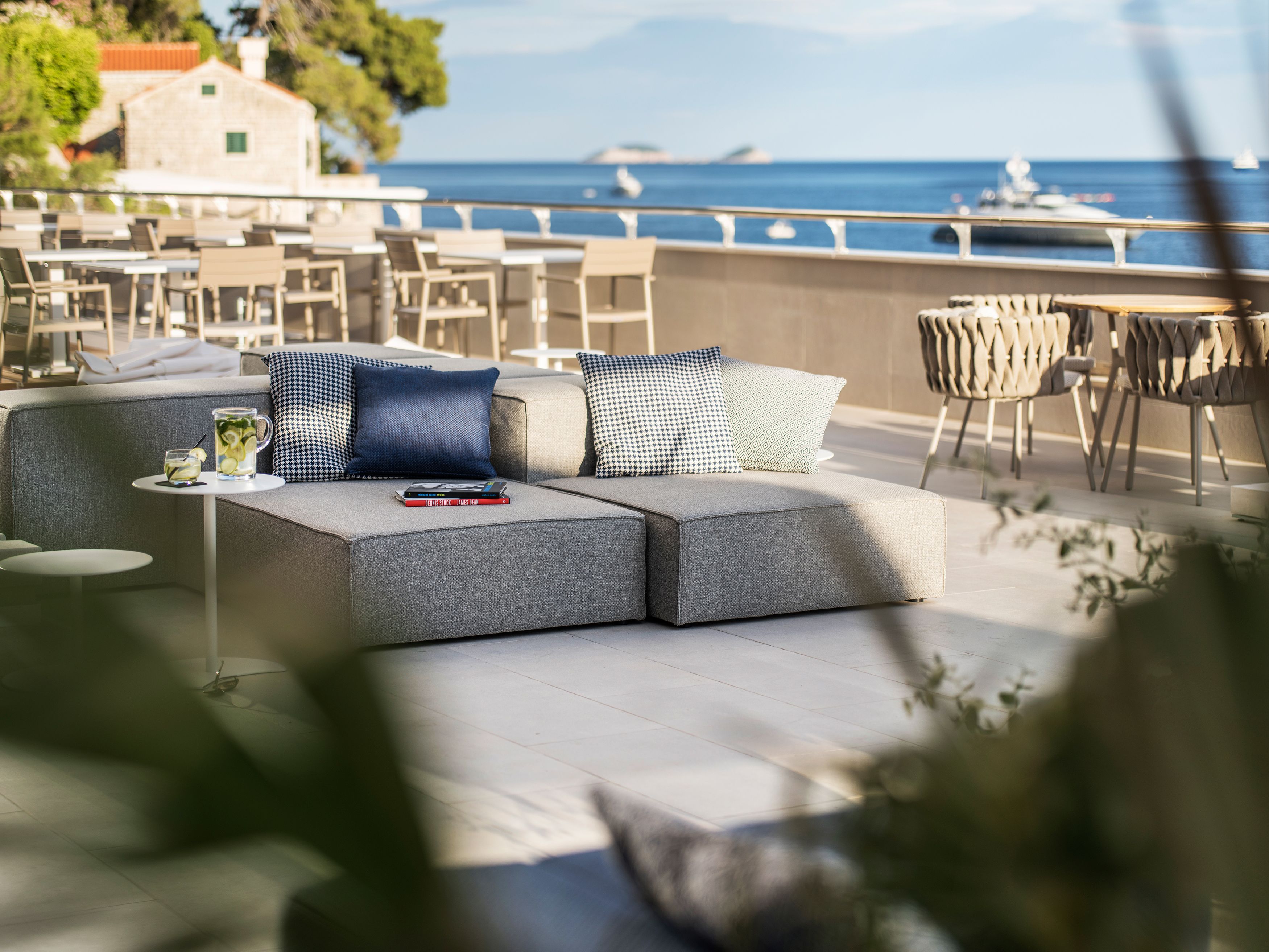 Un espace de détente extérieur moderne, avec un canapé et des fauteuils, une petite table sur laquelle se trouve une carafe de limonade, et une vue sur la mer et les bateaux au loin.