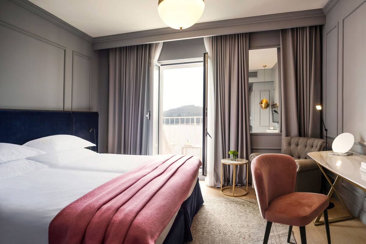 Quarto de hotel com uma cama grande, uma escrivaninha com cadeira, televisão e vista para uma varanda.