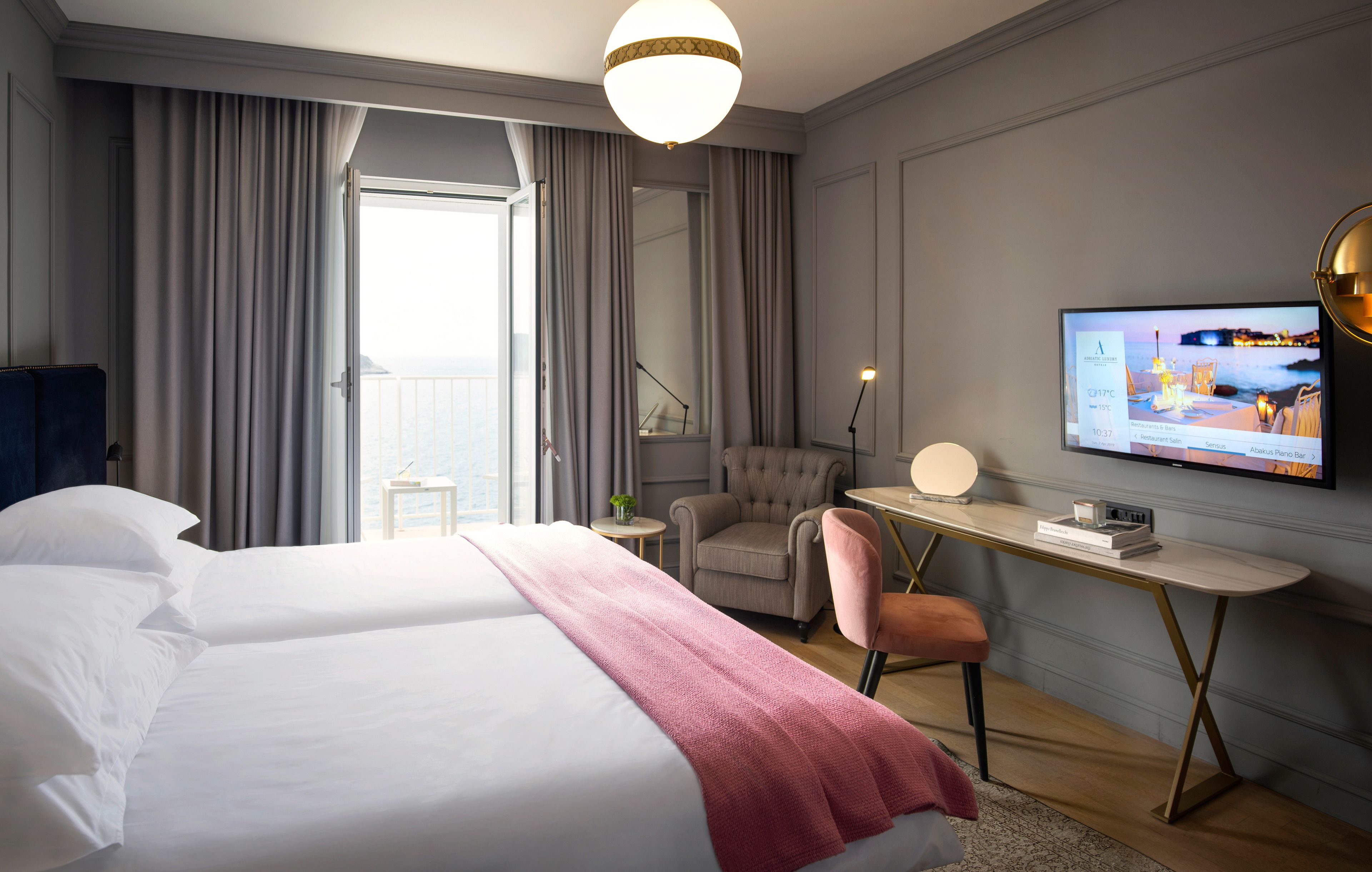 Ein Hotelzimmer mit einem großen Bett, einem Schreibtisch mit Computer, einem Sessel und einem Fernseher, der eine Speisekarte zeigt.