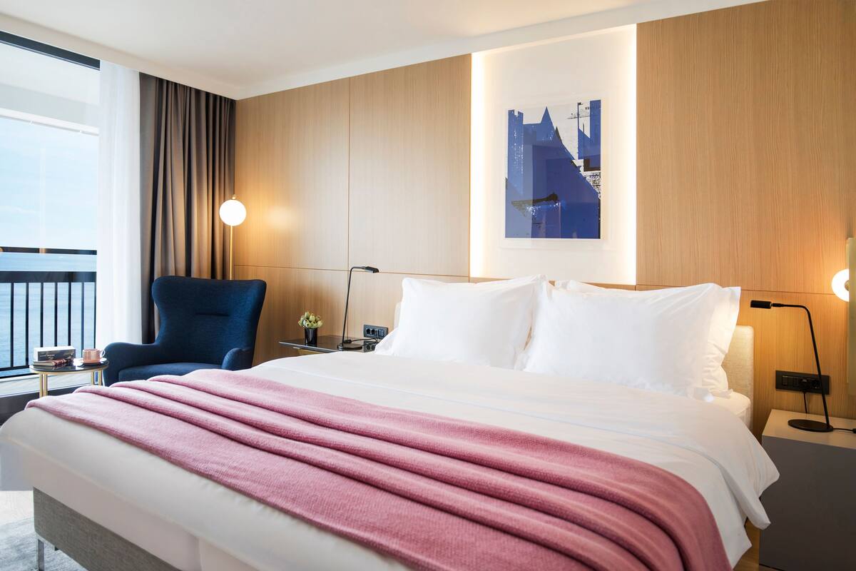 Quarto de hotel com uma cama grande, uma cadeira, uma escrivaninha, um abajur e um quadro na parede.