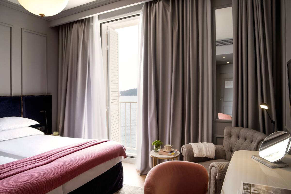 Quarto de hotel com cama, escrivaninha com luminária, cadeira e vista para o exterior.
