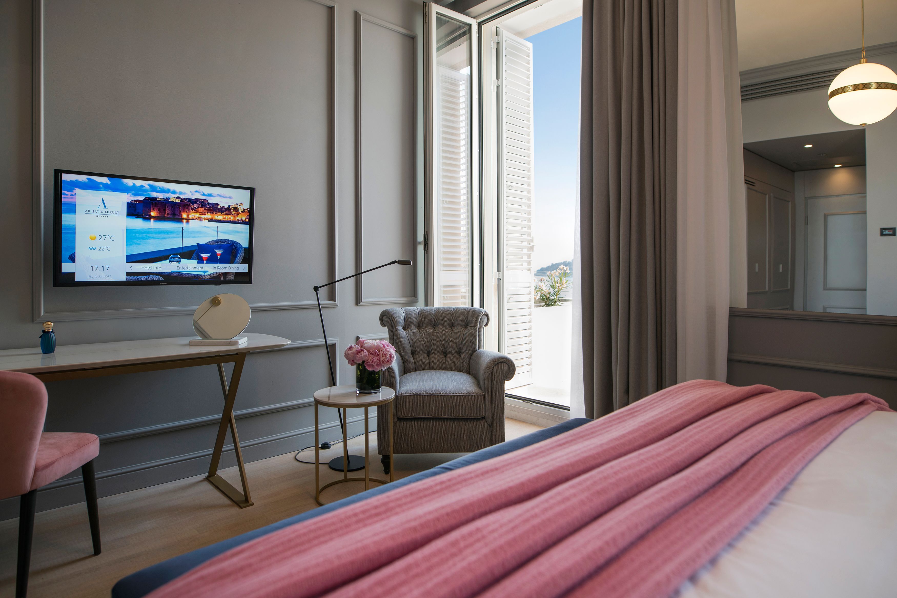 Ein Hotelzimmer mit Bett, Schreibtisch, Sessel und Fernseher, der Hotelinformationen zeigt, sowie Blick auf die Stadt durch das Fenster.