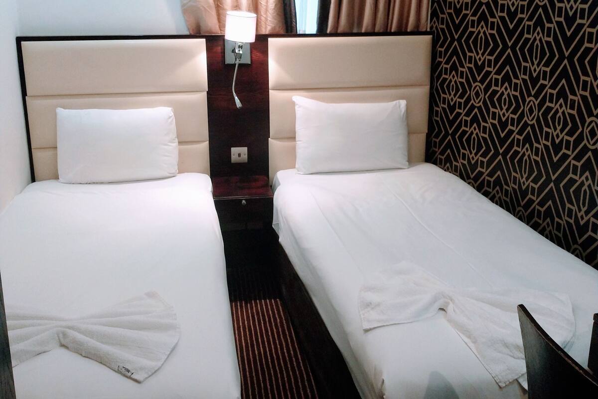 A hotel room with two single beds, a bedside table with a lamp, a patterned wall, and a window with curtains.
