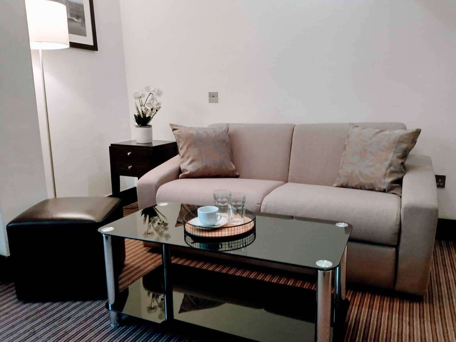 A modern hotel room with a beige sofa, a glass coffee table, a dark leather ottoman, a small side table with a plant, and a framed picture on the wall.