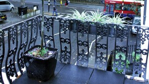 A balcony with a potted plant, a metal railing, and a view of a street with a red bus. - Edward Hotel Paddington (London)