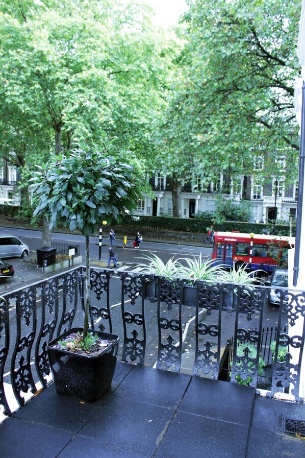Un balcon agrémenté d’un pot de fleurs, doté d’une rambarde en métal, et offrant une vue sur une rue où circule un autobus rouge.