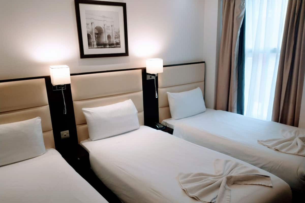 A hotel room with three beds, each with white linens and pillows. There are bedside lamps on either side of the beds. A framed picture hangs on the wall above the beds.