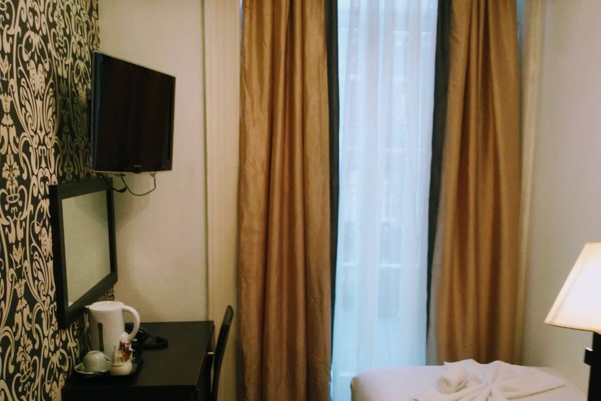 A hotel room with a bed, a desk with a chair, a television mounted on the wall, and a window with curtains.